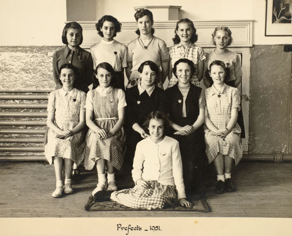 Eleven prefects, each wearing a badge, gather indoors in tiered rows for a group photograph, captioned "Prefects – 1951."