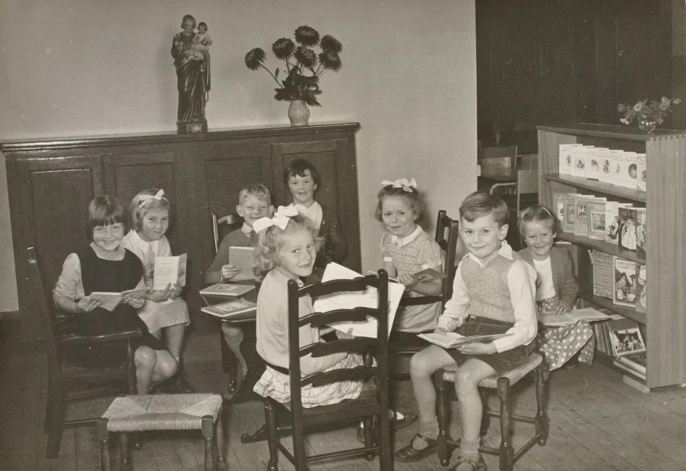 Young pupils sit reading picture books beside a bookcase, beneath a statue of St Joseph and Child Jesus on a cabinet.