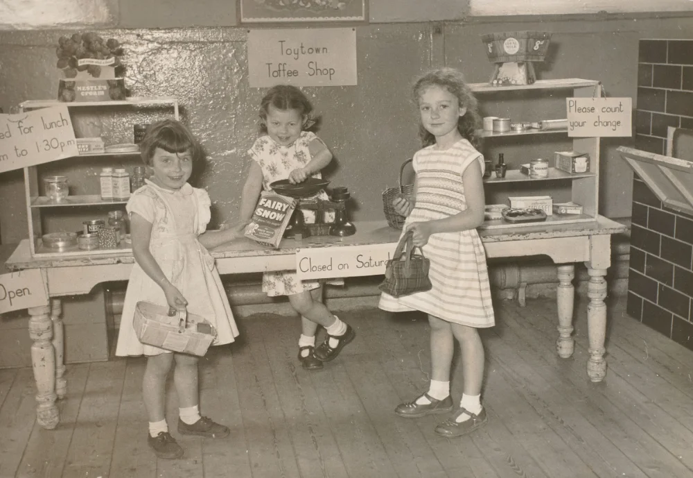 Three girls play shopkeeper and customer at a mock grocer's stall, "Toytown Toffee Shop," complete with weighing scales, tins and shopping baskets.