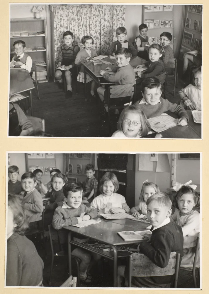 Infant pupils sit at classroom tables with books and papers, some holding pencils, near a bookcase and curtained window.
