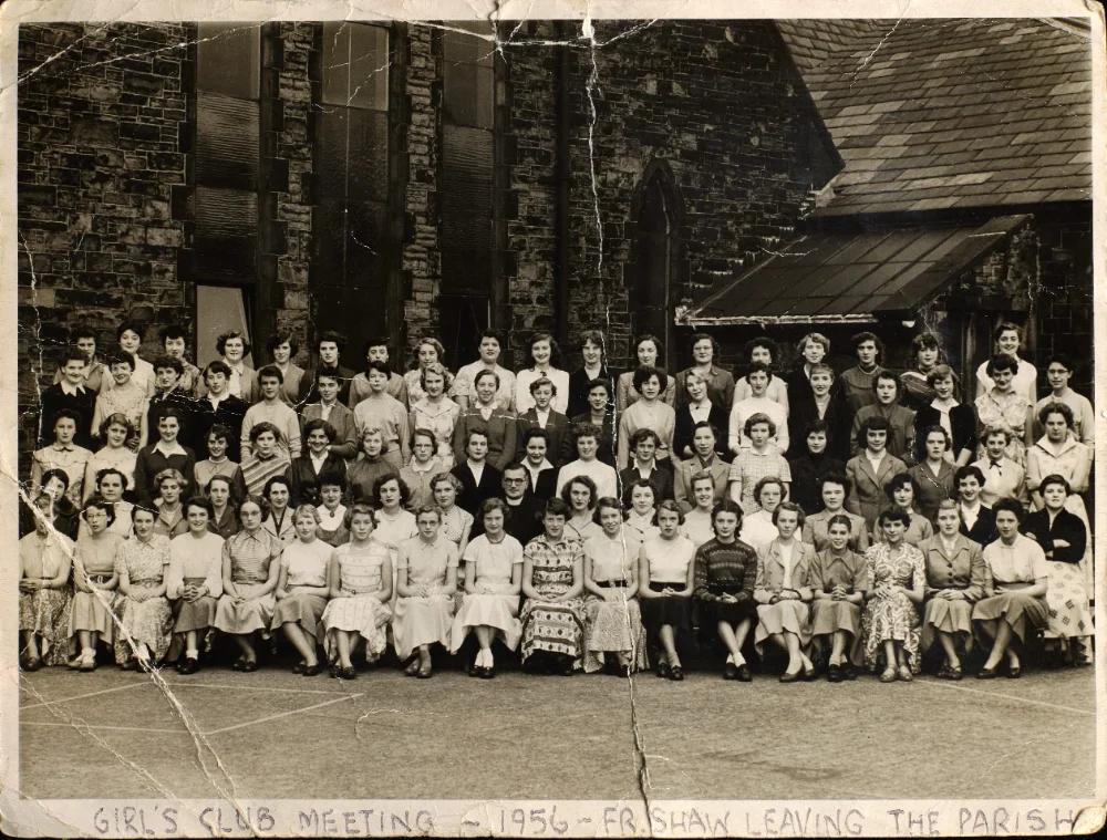 A large group of girls and young women gathers outside the church for a photograph, captioned "Girl's Club Meeting – 1956 – Fr Shaw Leaving the Parish."