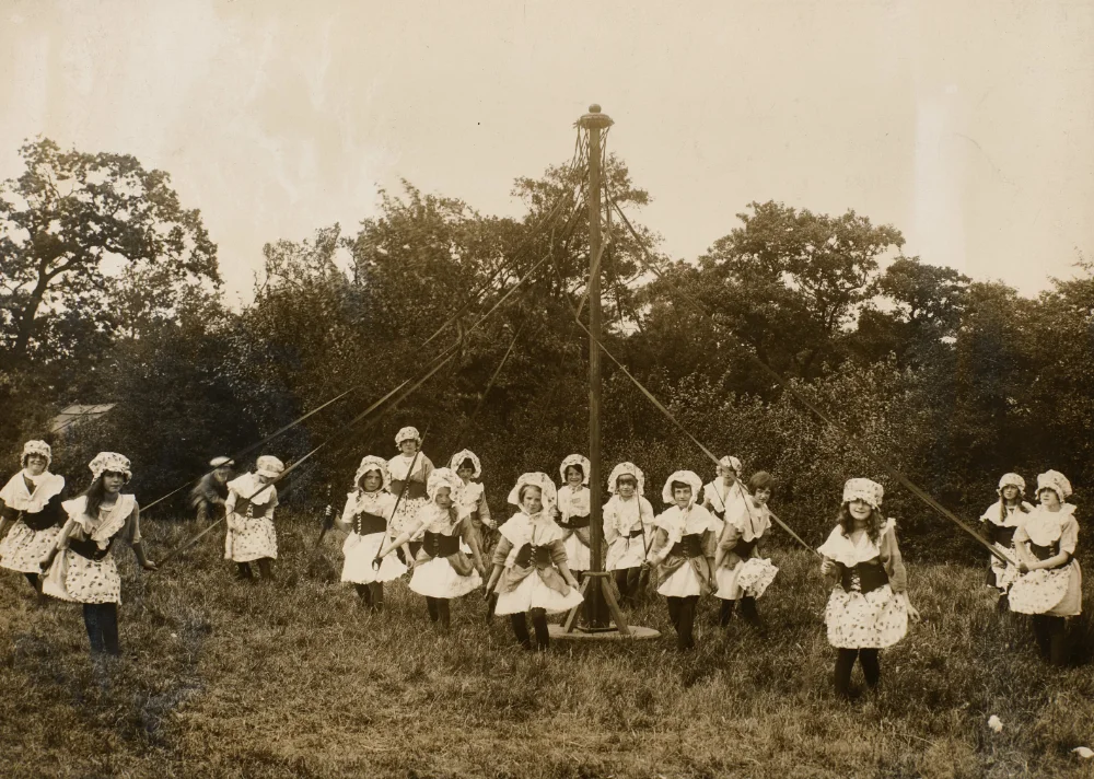Girls in matching bonnets, aprons and dresses hold ribbons around a maypole in a field, framed by trees.