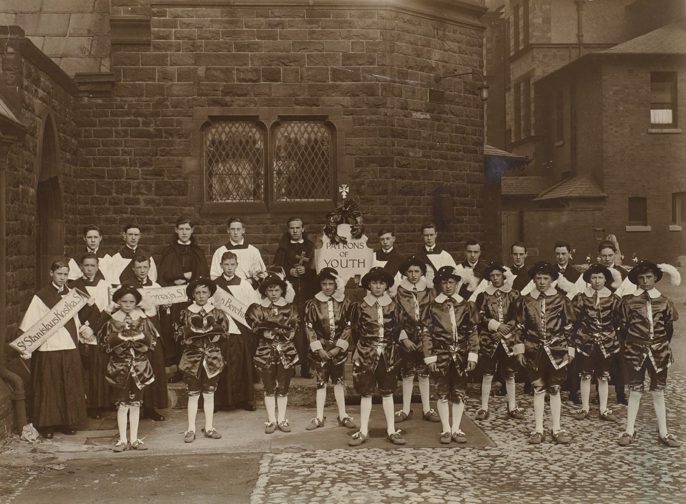 Boys dressed as patron saints of youth stand for a group photograph, altar servers and banners naming individual saints behind, a central banner reading "Patrons of Youth".