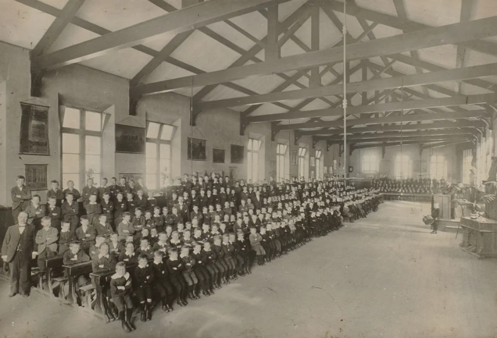 A large body of boys sits in long rows across the school hall beneath its timber roof trusses, wall maps and pictures hanging along the side.