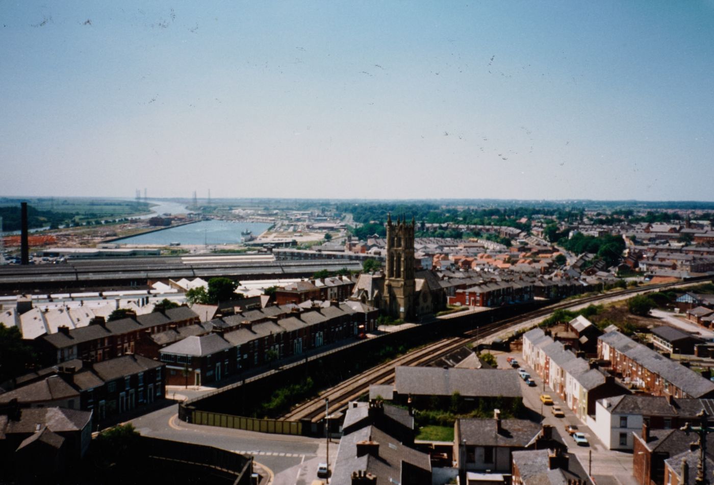 Colour photograph taken from height at St Walburge's, looking over terraced streets, railway lines and a neighbouring church tower toward the docks and river in the distance.