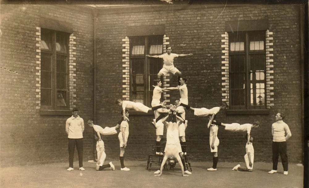 Boys in gym kit form a human pyramid in the schoolyard, watched by two instructors in tracksuits, a common school physical training exercise of the period.