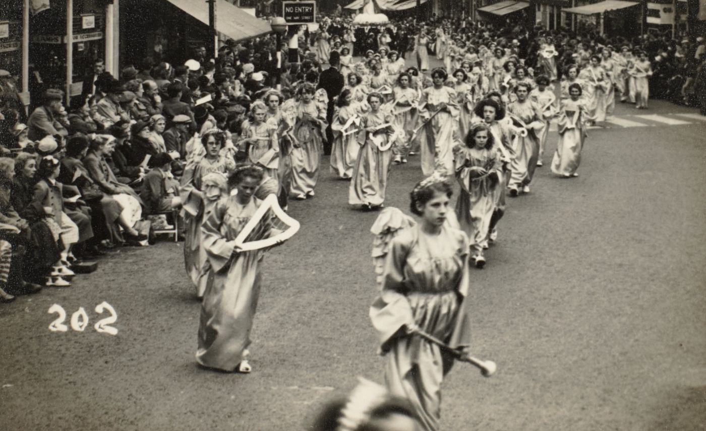 Girls dressed as angels with model harps and trumpets, processing down a Preston street, crowds seated along the pavement.