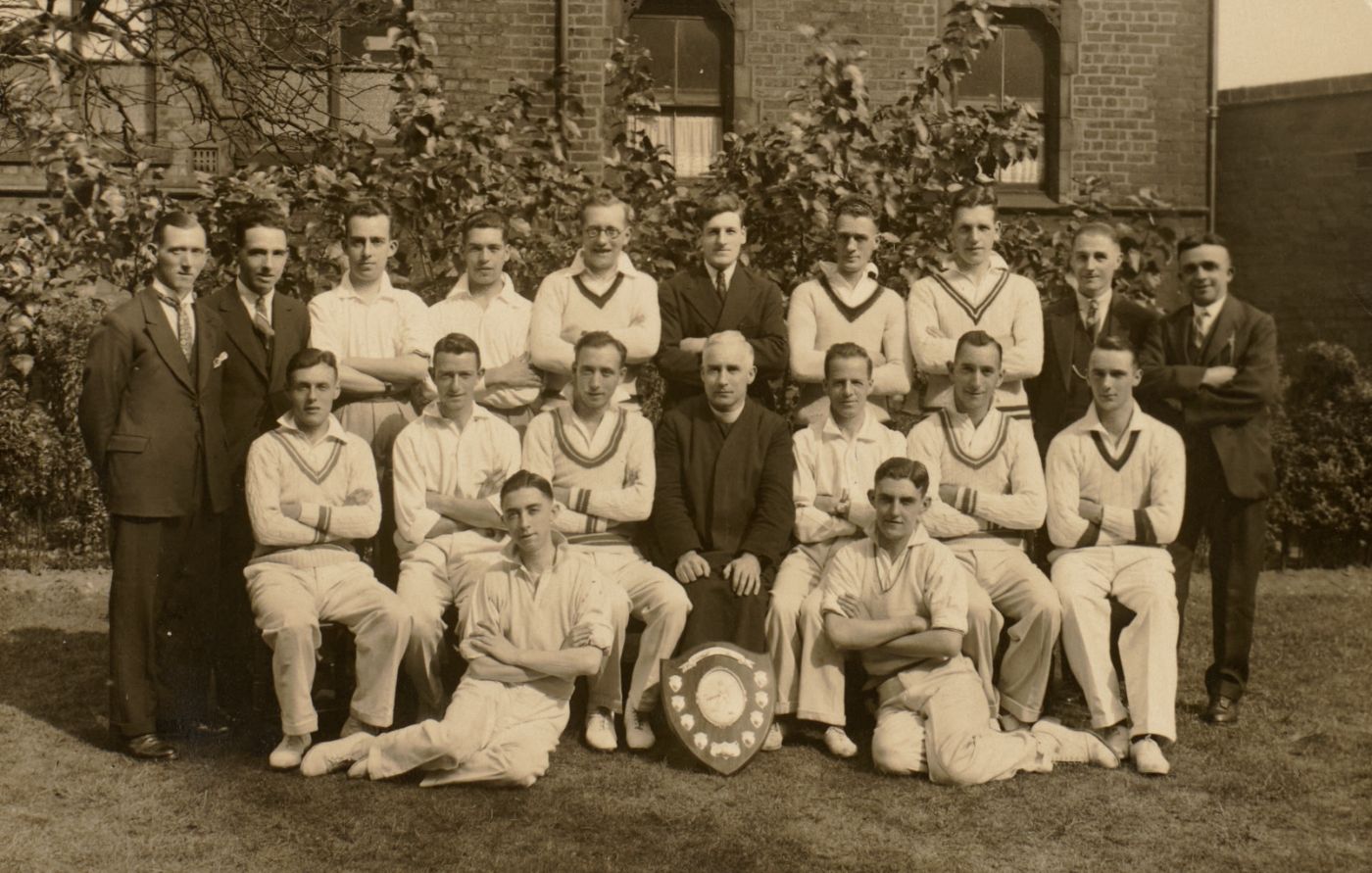 A cricket team in whites sits and stands for a team photograph outside a brick building, a priest seated centrally, a shield trophy displayed at the front.