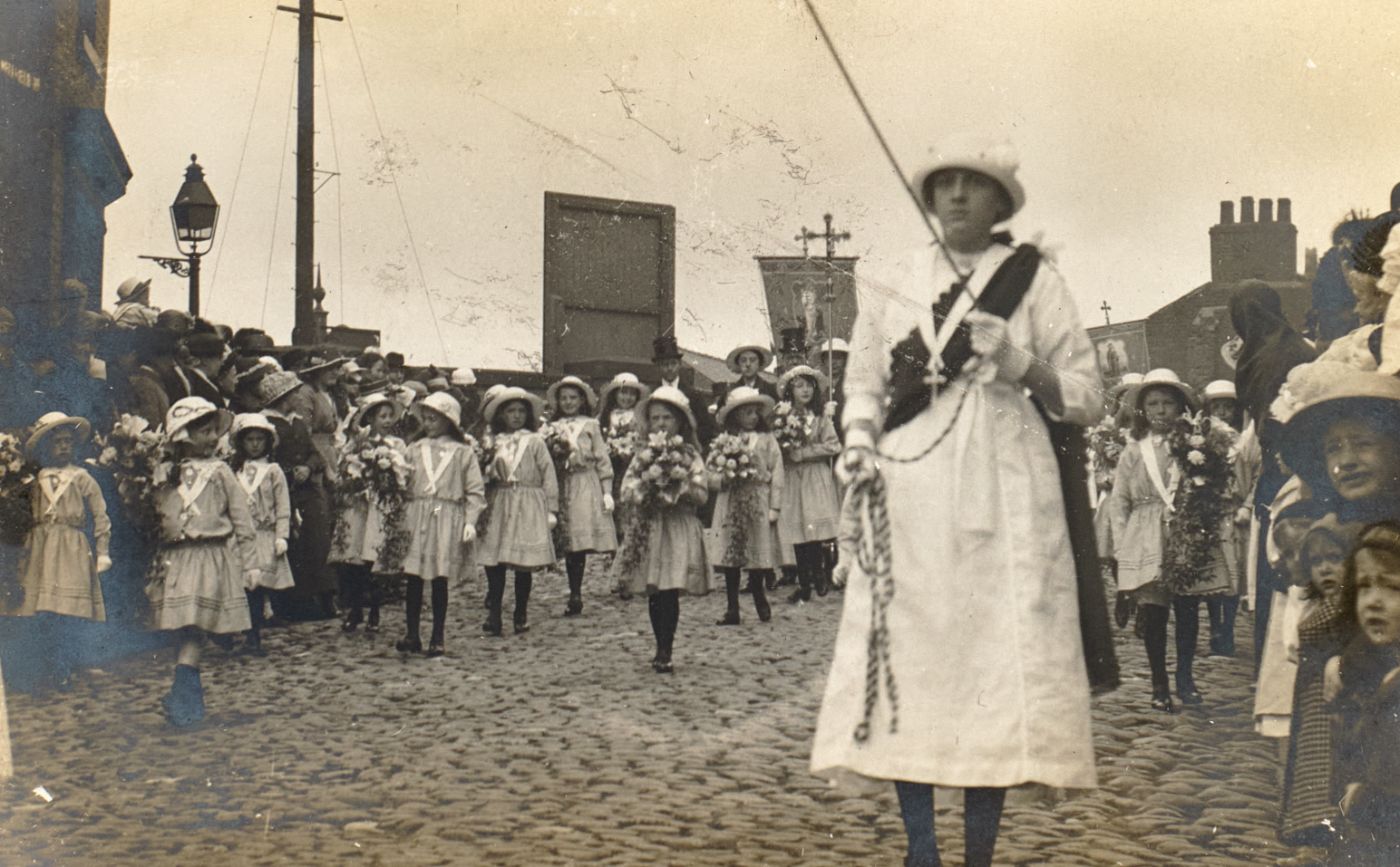 Girls in white dresses and sashes carry flowers along a cobbled Preston street, a woman in white leading with a processional cross behind, crowds watching from the pavement.