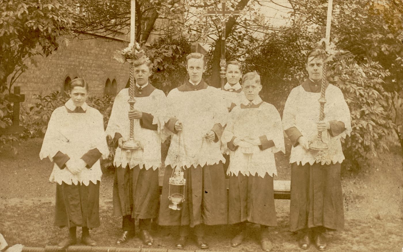 Altar servers in cassock and cotta with a processional cross, candlesticks and thurible, standing in the churchyard.