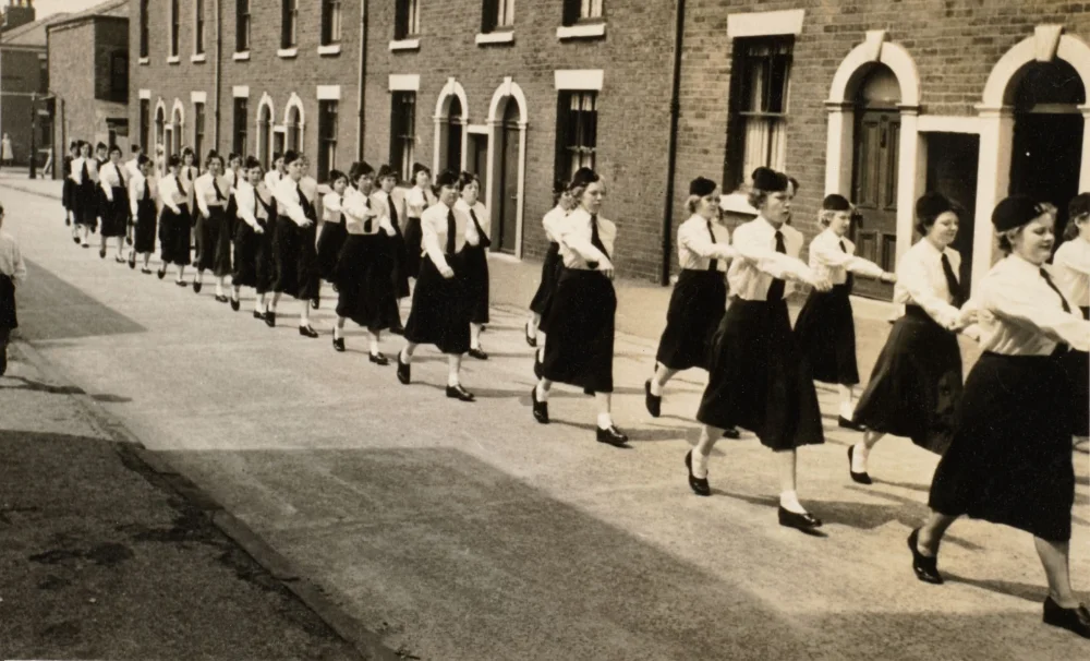 A long line of girls in matching berets, ties and dark skirts marches arm in arm along a residential street lined with terraced houses.