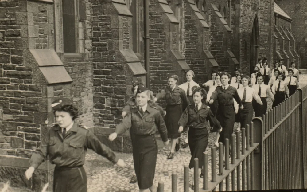 A long line of girls in uniform, some in tunics and berets, marches arm in arm past a stone building within the church grounds.