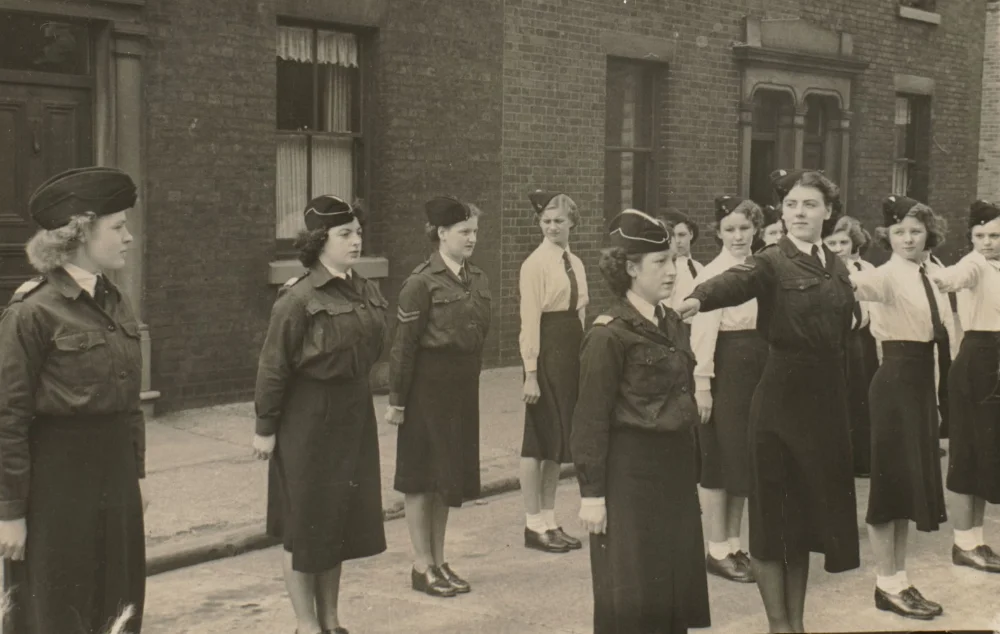 Girls in uniform stand in ranks outside a terraced building, one with arm extended as if directing drill positions.