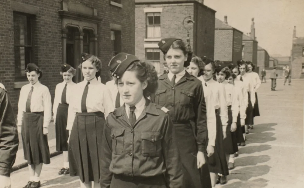 Girls in uniform, some with peaked forage caps, stand in a long line along a residential street.