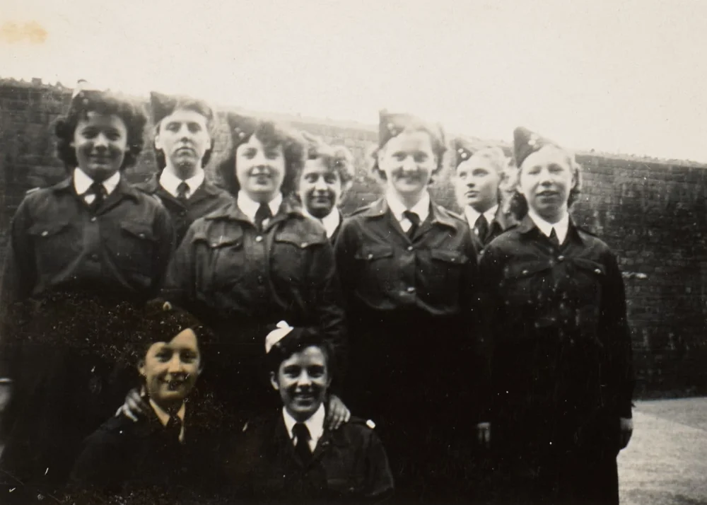 Nine girls in tunic uniforms and forage caps gather in two rows before a brick wall for a group photograph.