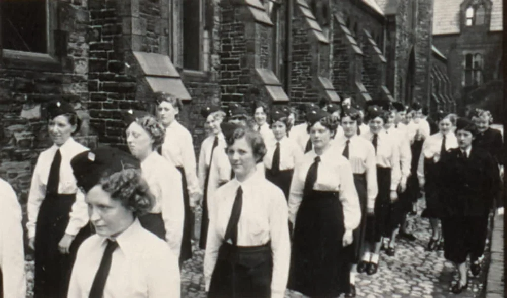 Girls in uniform with ties and forage caps march in rows past a stone building within the church grounds.