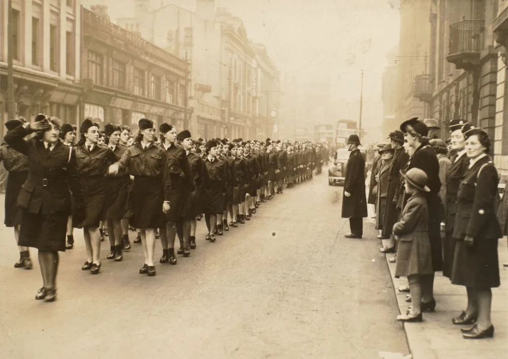 Long ranks of girls in uniform march down a Preston street past a police officer, spectators and salutes from the front row, a double-decker bus visible ahead.