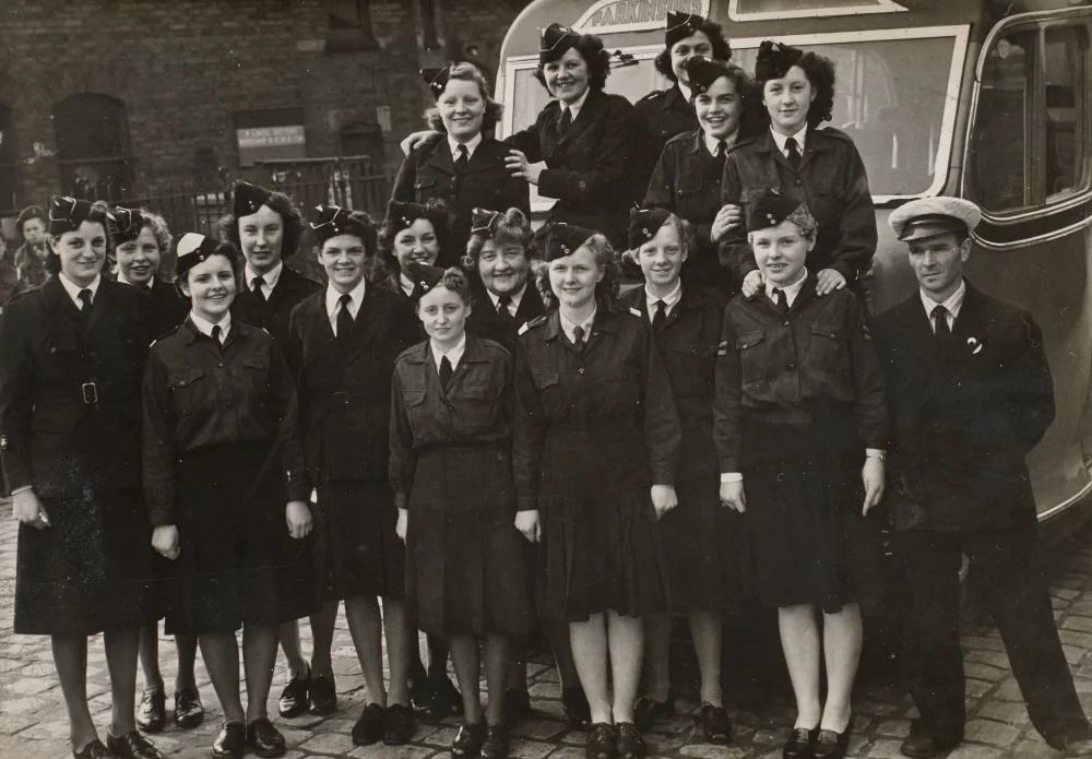 Women in dark military-style uniforms and peaked side caps, with others in lighter shirts and jackets, stand in rows beside a bus outside a brick building. Male driver at right.