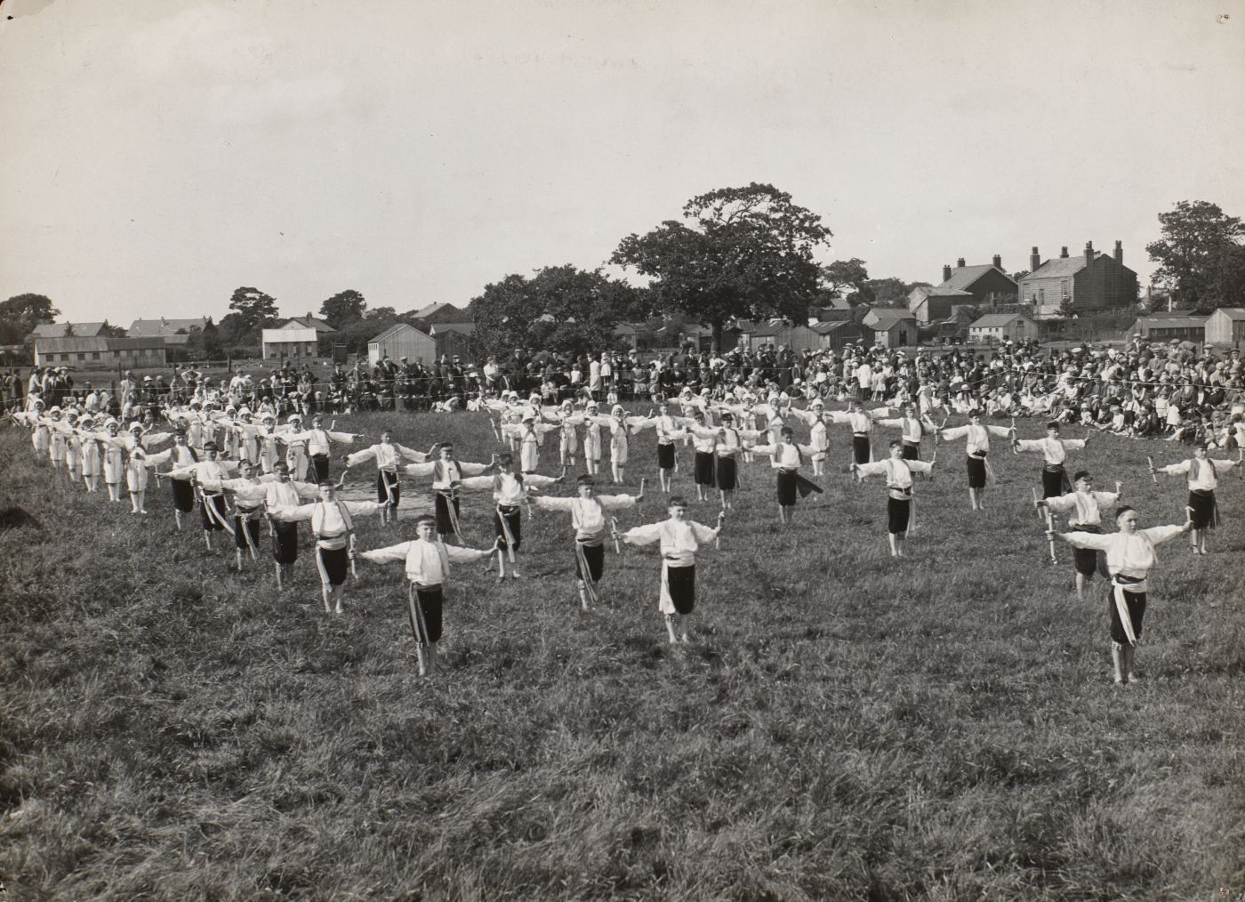 Rows of boys in matching drill uniform perform synchronised exercises in a field, a large crowd of parents and parishioners watching behind a rope boundary.