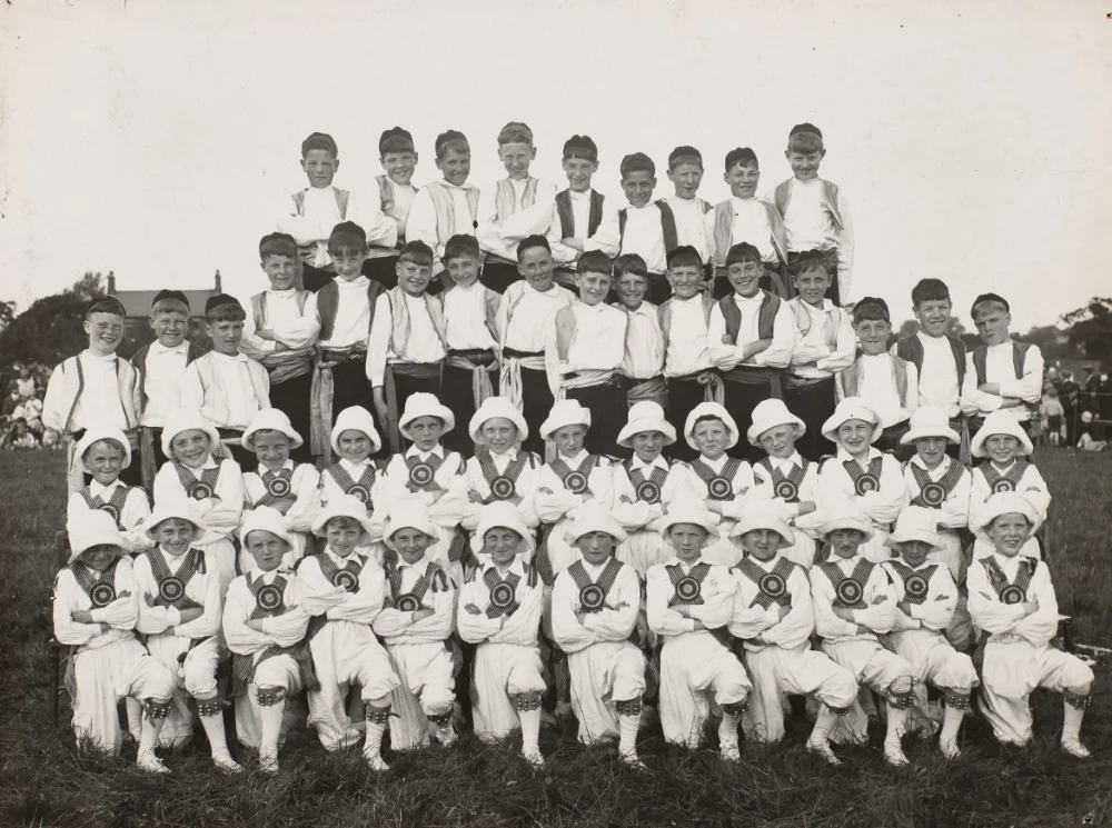 Boys in matching sashes, waistcoats and decorated tunics, some wearing round badges, gather in tiered rows on grass, likely for a school fête or sports day performance.