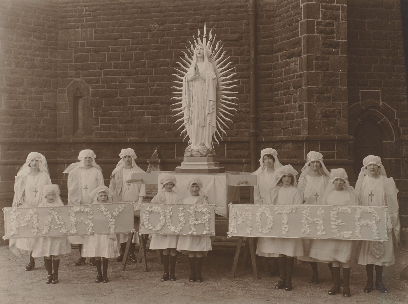 Girls and women in white veils pose beside a statue of Our Lady, holding flower-decorated banners reading "Mary Our Mother."