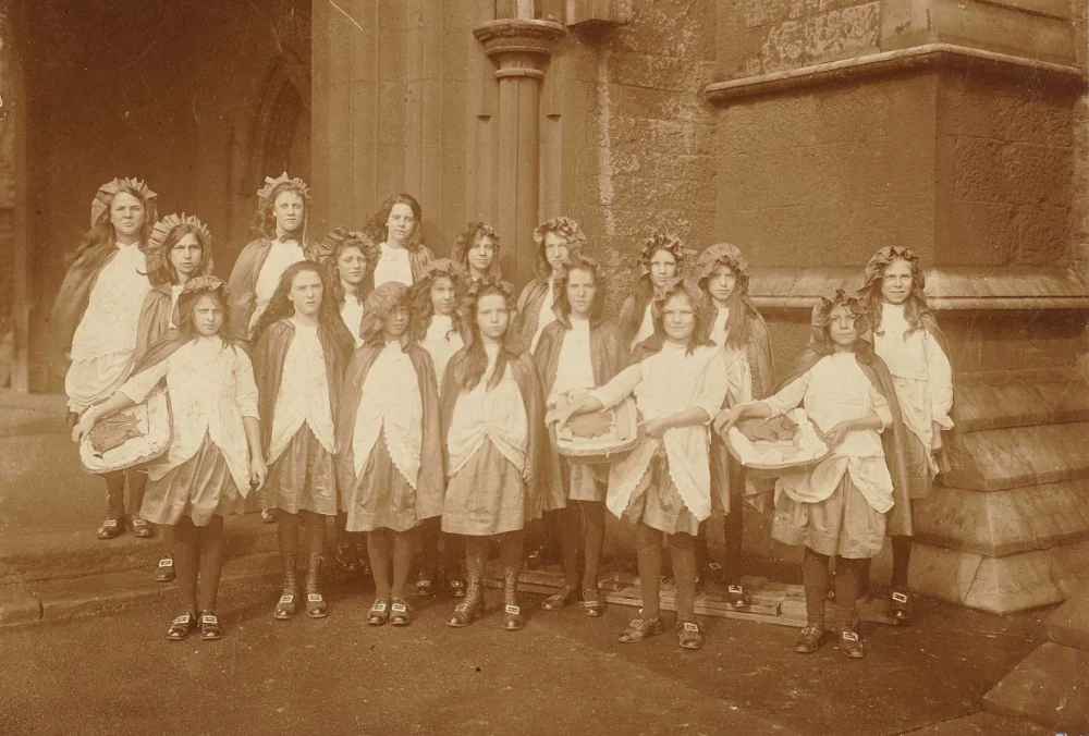 Girls in mob caps and aprons stand for a group photograph outside the church.