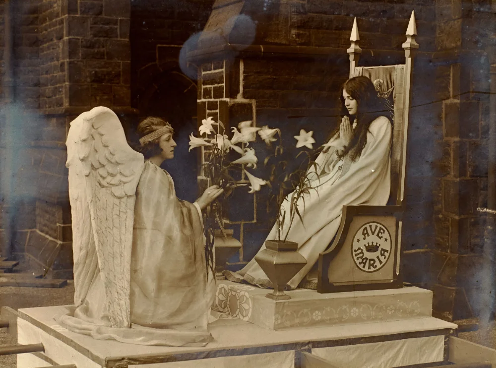 A scene of the Virgin Mary and the Angel Gabriel offering lilies, staged outside a church on a platform inscribed "Ave Maria."