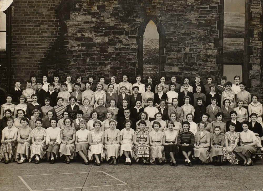 A large group of young women and girls, arranged in four rows, gather in front of a stone church wall with a pointed arch window.